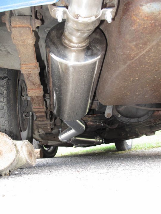 Supercharged Pistonheads - The image shows an undercarriage view of a vehicle, likely a car. The focus is on the muffler, which appears to be made of stainless steel and is attached by pipe and clamp to other undercarriage components. The muffler is positioned between the vehicle's components, as a part of the engine exhaust system. The muffler has a smooth, somewhat cylindrical shape that narrows slightly toward one end. In the background, a partially visible dampener can be seen, which is standard in cars to reduce road noise.