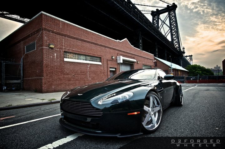 Car Porn - Page 1 - Aston Martin - PistonHeads - The image shows a sleek black Aston Martin sports car parked on the street. The car has a design packed with character, featuring distinctive wheel rims, a large front grille, and a side profile that suggests both luxury and performance. Behind the car, there is a brick building, and in the background, a suspension bridge can be seen. The sky overhead is cloudy, suggesting either early morning or late afternoon, times that often provide the most dramatic lighting conditions for photography. The overall setting suggests an urban environment, possibly near the waterfront given the presence of the bridge.