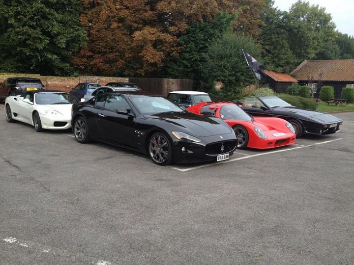 A car is parked on the side of the road - Pistonheads - The image shows a parking lot filled with luxury cars. These cars are designed to look futuristic, with the front end of a sports car and the body shape of a retro vehicle. The cars are parked in a diagonal manner, and they are all black, with a glossy finish reflecting the ambient light. The setting appears to be a suburban or semi-urban area, with trees visible in the background. The ground is gray and asphalt appears wet or has recently been rained on, indicating that the photo may have been taken after a recent rainfall.