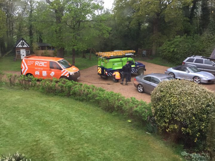 Super Car Sunday - Page 1 - Goodwood Events - PistonHeads - The image shows a parking area with an orange van on the left parked beside a green utility truck with a large ladder mounted on top. Two individuals are standing in front of the truck, seemingly engaged in a conversation or inspecting it. On the right side of the image, there is a line of vehicles including cars and trucks parked on a gravel surface beside a hedge and bushes. The setting appears to be an outdoor, possibly rural area with greenery and trees in the background.