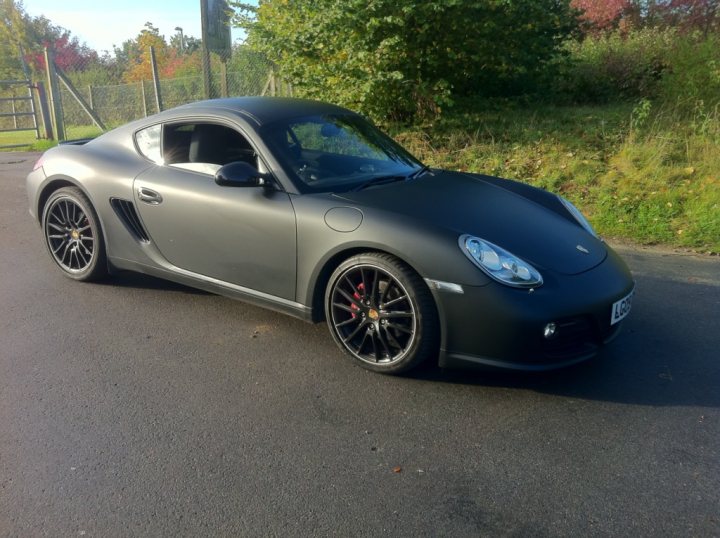 Loves Hire Pistonheads - In the image, a black Porsche Cayman sports car is parked on a tarmac road. It features black rims and alloy wheels, adding to its sleek design. The car is positioned against a backdrop of lush greenery, with a tree and a fence visible in the area. The scene is bathed in natural light, with the sky forming a serene blue backdrop. The overall image showcases the car's beauty and the tranquil setting it's parked in.