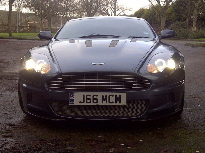 DBS wheels on DB9 - (more or less) - Page 1 - Aston Martin - PistonHeads - The image captures a moment on a wet pavement under an overcast sky. A sleek, black Aston Martin sports car, adorned with a rectangular registration plate that reads "J66 MCM", is the main focus of the image. The car's stylish grille and headlights glisten with reflections from the wet road. The background offers a glimpse of a serene park filled with trees and a grassy lawn. Despite the detached tire in the background, the overall scene exudes a sense of elegance and luxury.