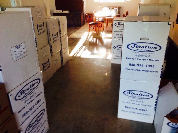 A white refrigerator freezer sitting inside of a kitchen - The image depicts a room that appears to be in the process of moving or reorganizing. The main focus is a large, open space that looks initially cluttered. The floor is covered in various color palettes and patterns, indicating the presence of several boxes stacked around the room. In the center, there's a cardboard box with some blue writing on it, possibly indicating a brand or company like 'Stration'. Scattered throughout the room are several other boxes and some chairs, suggesting recent activity. The room is bright and well-lit, with natural light coming in from a window on the right. The presence of a counter area with a table and chairs in the background suggests that the space may serve multiple purposes.