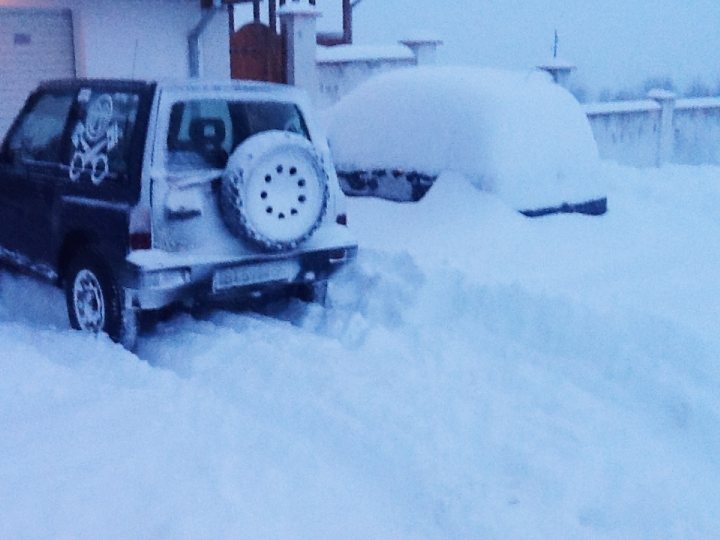 Pics of your offroaders... - Page 13 - Off Road - PistonHeads - The image depicts a snowy scene featuring a parking spot covered in deep snow. A black SUV, which seems to have been covered in snow as well, is partially buried under a thick layer of snow, with only its front section visible. Behind the SUV, there is a white vehicle also covered in snow, suggesting recent heavy snowfall. The white SUV's rear wheel, recognizable due to its distinctive black tire with silver rims, is sticking out from the snow, which has settled more heavily around the other wheels. The background is dominated by a white house with a fence, adding a sense of urban domesticity to the overall scene. The photo is taken in a low-angle perspective, giving prominence to the vehicles and framing the sky above them.