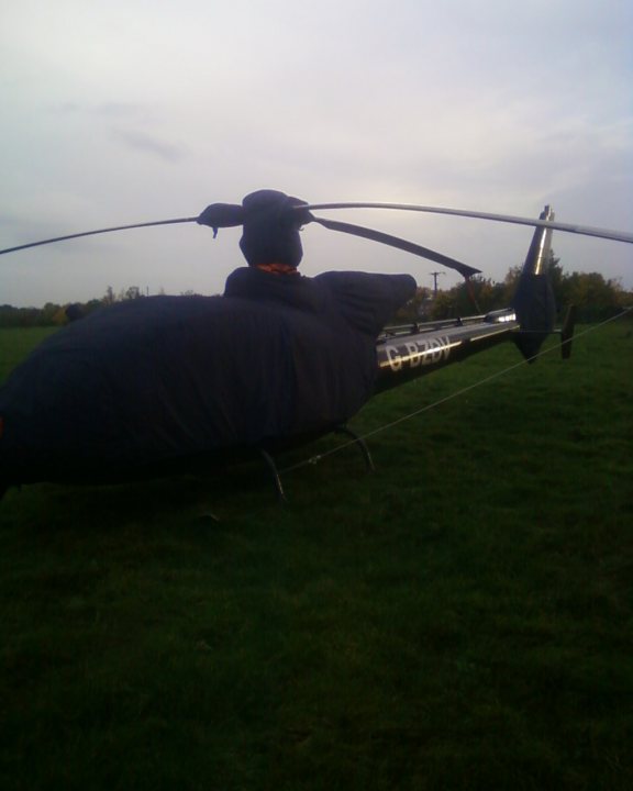 Helicopter Gazelle Pistonheads Fancy - This is a photorealistic image showing a large, dark-colored inflatable object, possibly a balloon or an artificial weather doppler, sitting on a green grassy field. The inflatable is characterized by a black body with a gold or silver section near the base and a curved neck extending into the air. The neck appears to be tethered or anchored to the ground with a visible cable or wire. This object is quite hairy, suggesting that the tether may be interacting with it in a way that obscures other features. The photo's quality is somewhat rough, contributing to the blur and overall lack of distinct detail in the scene.