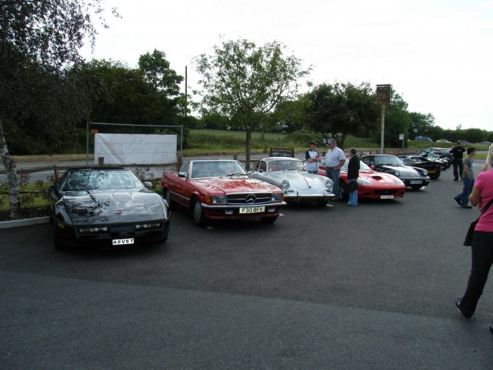 Breakfast Meeting, The Hare, Roxwell 4th June - Page 1 - Kent & Essex - PistonHeads - In the image, a group of people are gathered in a parking lot, engrossed in admiring several vintage cars. The cars are lined up neatly, their glossy bodies reflecting the daylight. The most prominent of these vehicles is a red vintage Mercedes, its vibrant color contrasting with the blacktop. Behind the cars, a backdrop of trees under a gray, cloudy sky adds a serene touch to the scene. The spectators, some of whom are holding handbags, appear to be absorbed in the classic elegance of the cars on display.