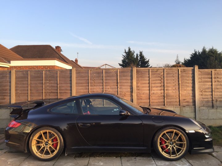 Weiss gold wheels - Page 2 - 911/Carrera GT - PistonHeads - The image shows a black Porsche sports car parked on a paved area outside a residential building. The car's body has unique gold-orange alloys which contrast with its dark color. The car features a large rear wing, likely for added downforce, and it appears to be a convertible model due to the visible soft top. The setting sun casts a warm light on the scene, revealing a wooden fence and the silhouette of pine trees in the background.