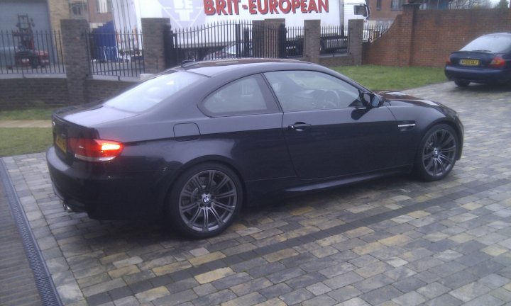 Used E92 M3 DCT Pricing? - Page 3 - M Power - PistonHeads - This image features a black BMW car parked on a cobblestone driveway. The car is positioned either in front of or parked next to two parked white delivery vans. A brick or block wall is also visible behind the vehicle. The driveway itself appears to be a mix of brickwork and possibly larger stones, giving the area a rustic look. The photo seems to capture a calm, stationary moment, perhaps suggesting the onset of a delivery or loading process with the presence of the vehicles.