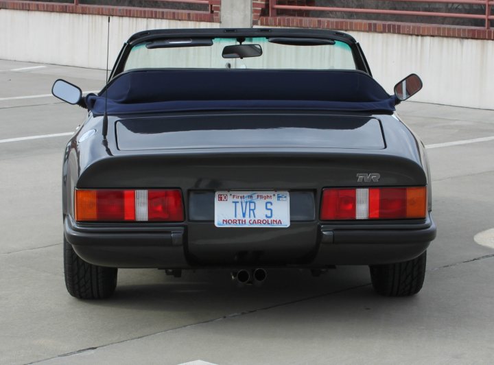 License plate on a TVR S - Page 1 - TVR in USA - PistonHeads - The image shows a black sports car parked in a white-stripe parking lot. The vehicle is angled so that it is not fully visible from this perspective, but the rear view of the car and its two-tone tail lights are clearly displayed. The North Carolina license plate is visible on the car's trunk. The car has the windows down on both sides, and there is a sundress draped over the back seat, catching the breeze. The background features an outdoor structure that implies the setting is possibly an urban or public park area.