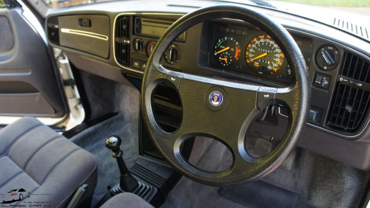 Classic Saab 900 26000 Miles - Page 1 - Saab - PistonHeads - The image shows an interior view of a car, focusing on the steering wheel and instrument panel. The steering wheel is covered in black leather trim, with symbols and markings visible on its surface. Behind the steering wheel, the dashboard displays various gauges, including a speedometer, fuel gauge, and a clock. There's a logo of a silver car brand visible in the middle of the dashboard. The car appears to be an older model, with a classic design. The image captures the condition of the car at a moment of stillness, with the seats appearing unoccupied.