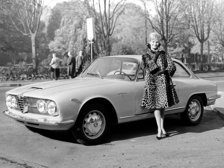 Alfa Romeo Sprint 2600 Bertone  - Page 1 - Classic Cars and Yesterday's Heroes - PistonHeads - The image is a vintage black and white photograph. It features a woman in a leopard print coat and a fur stole, leaning against an Alfa Romeo car. The woman is positioned in the center of the photograph, flanked by two men standing further back. The car has distinctive designs, exhibiting a compact and classic exterior. They are all situated in a paved area with trees in the background.