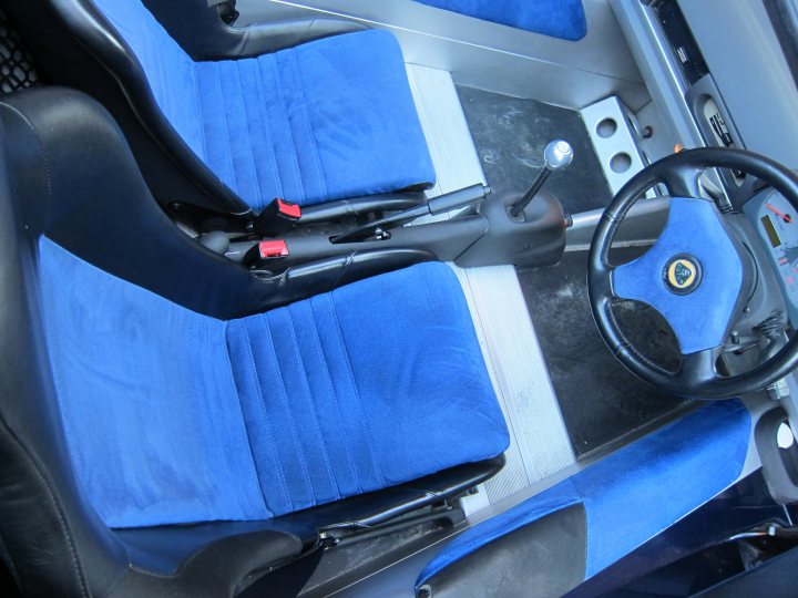 Elise S1 Millennium Edition, 1 owner from new... - Page 1 - Readers' Cars - PistonHeads - The image shows the interior of a vehicle from the perspective of the front passenger seat. The seating area is fitted with blue leather upholstery, which is typical for higher end vehicles. Visible on the left seat cushion is a small black tag. The steering wheel is on the right side of the image, which indicates that this vehicle has a right-hand drive configuration, likely for use in Europe, Asia, or Australia. The visible portion of the dashboard and the steering wheel hub shows some controls and labels, but the specific brand or model is not clearly identifiable. There appears to be some grain on the surface of the seating, possibly indicating the texture of the leather material or a pattern design.