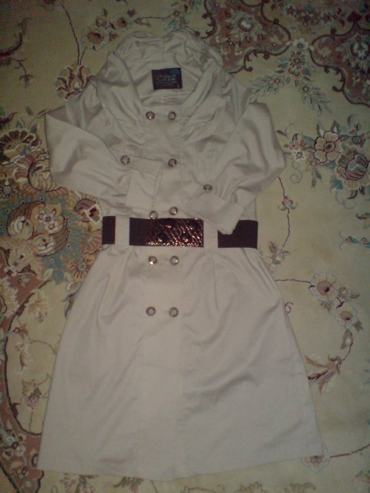 The image shows an elegant dress laid out on a patterned fabric, with the focus on the upper back area. The dress appears to be white, with three visible buttons at the center-back of the garment. The fabric is adorned with a shiny black belt with a buckle, which adds a charming contrast to the design. The background is blurred, emphasizing the dress as the main subject of the image. The overall style of the image is simple and clear, with the dress being the star of the show.