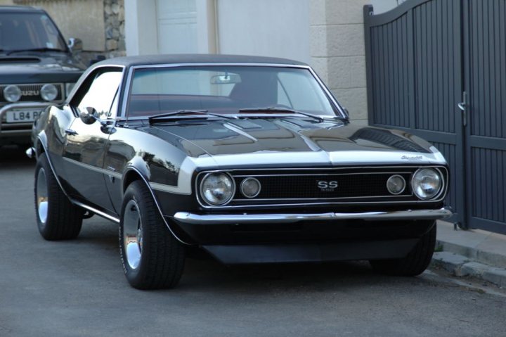 Camaro Pistonheads Worth - This is an image of a black classic car, likely a Ford Mustang, captured on a street. The photo is taken in natural light, and the car appears to be a convertible model, given the visible soft top. Two other vehicles, possibly cars as well, can be seen in the background, with traffic lights partially visible, indicating the car is paused at an intersection. The car has silver trim and a distinctive grill.