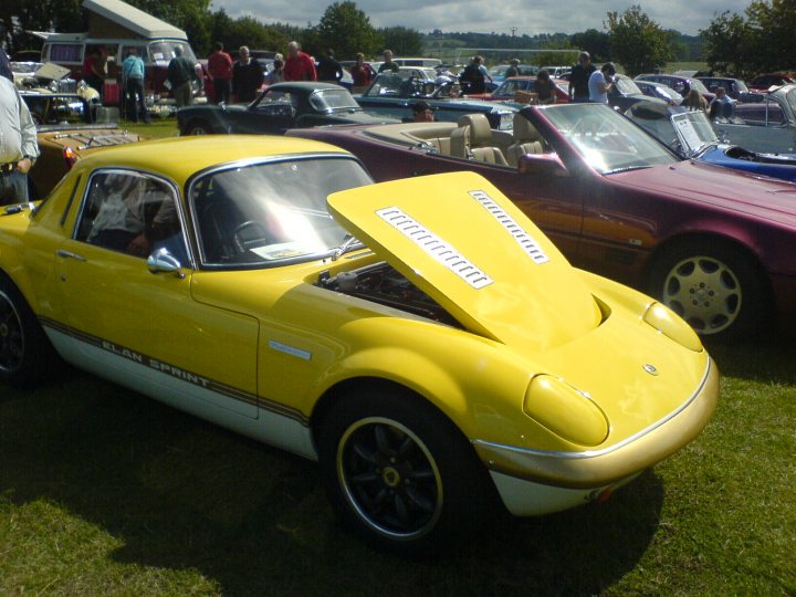 Pistonheads - The image depicts a vibrant scene at a car show. The main focus is on a vintage yellow sports car, which has had its top removed and now features a bright yellow convertible hood. There's an array of other vintage cars parked in the background, including a purple and black one right next to the yellow one. The cars are lined up on what appears to be a grassy field, suggesting a casual, outdoor event. Several people are scattered around the area, possibly viewers or participants of the show. The overall atmosphere appears lively and enthusiastic about vintage automobiles.