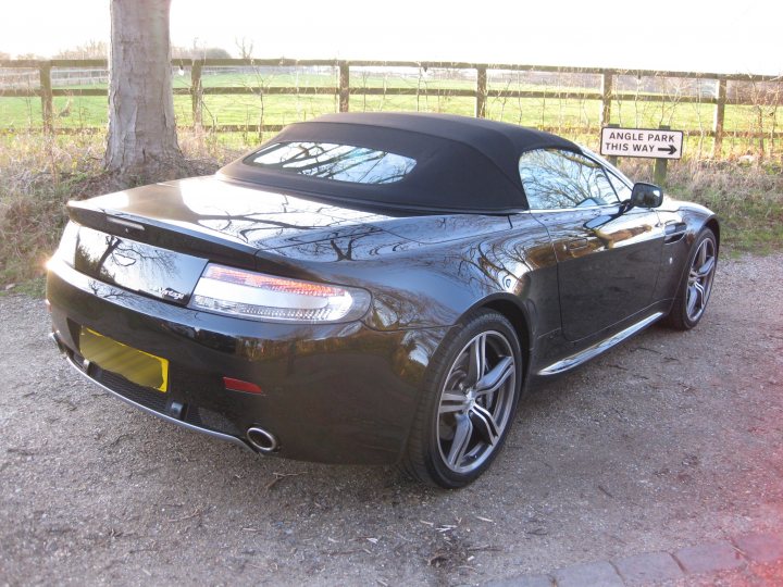 Back in the fold and its feeling good!!!! - Page 1 - Aston Martin - PistonHeads - This image depicts a sleek, black convertible car parked outdoors. The car is parked on a gravel surface with patches of grass visible. It is equipped with alloy wheels and a retractable hardtop that is currently down, making the car a convertible. Yellow and black license plates are visible at the back of the car, indicating its registration. The setting appears to be a rural area with fencing and a sign behind the car. The sunlight suggests it might be on the heels of sunrise or late afternoon judging by the warm tones with light reflected off the car's surface.