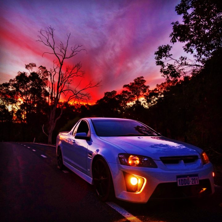 Ute Numbers in the UK??  - Page 1 - HSV & Monaro - PistonHeads - The image captures a breathtaking scene at sunset. A sleek white car is parked on the side of a winding road, which seems to be made of asphalt. The backdrop is a stunning display of colors from the sunset, casting soft hues on the horizon. The sky is painted with vibrant shades of orange, yellow, and pink, creating a picturesque view. The road is flanked by trees on both sides, adding a touch of green to the otherwise warm palette. The car's interior light is on, illuminating its presence in the serene and colorful landscape.