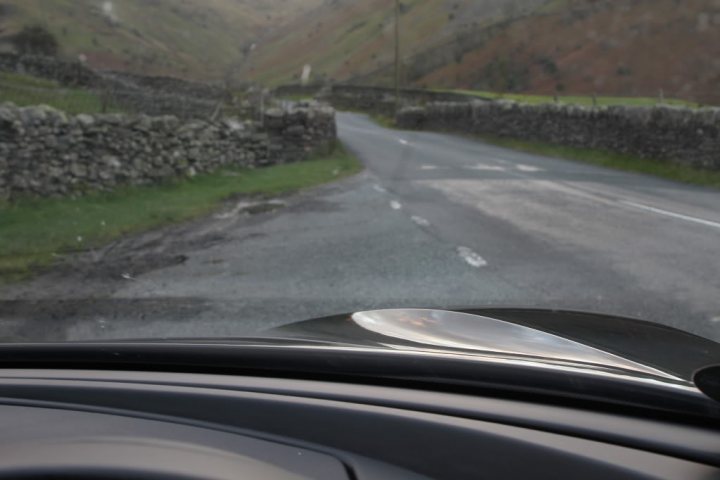 black evora spotted - Page 1 - Evora - PistonHeads - The image displays the front view of a car dashboard through the windshield. The car seems to be in motion, as indicated by the motion blur of the road ahead. The road appears to be a rural, one-way street, with a rock wall on the left side and a grassy area on the right. There's a stone wall lining the road, suggesting a countryside setting. The colors in the image are muted, with earth tones dominating the scene.