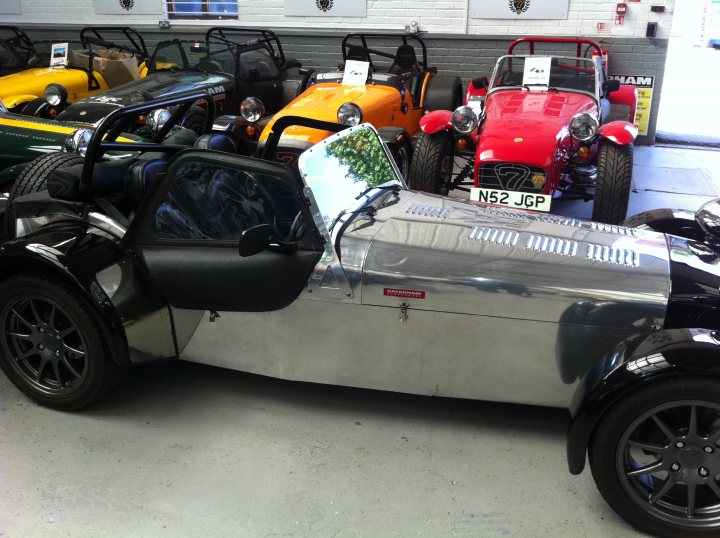 I've gone ahead and done it. - Page 1 - Caterham - PistonHeads - The image presents a vibrant display of vintage cars and motorcycles, parked neatly in rows inside a well-lit garage. The garage's interior walls are brick-like, lending a rustic charm to the scene. In the foreground, a unique car stands out, its elongated body resembling that of a custom-made convertible with its side windows open. The vehicles, including models with sleek design elements, suggest a collector's or enthusiast's private collection. The overall scene conveys a sense of nostalgia and appreciation for the elegance and craftsmanship of these classic vehicles.