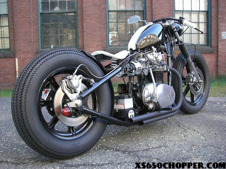 Building a Bobber motorcycle - Page 1 - Biker Banter - PistonHeads - The image features a custom-built motorcycle, prominently styled with large, knife-edged guards that give it a menacing appearance. The bike's design elements, such as the heavily-modified saddlebags, detailing around the wheels, and the faux-spike bodywork, suggest that it has been heavily customized. The motorcycle is equipped with a substantial rear wheel drive system. In the background, there is a partially obscured brick building with partial windows. The image appears to be taken outdoors during the day on a sunny day.