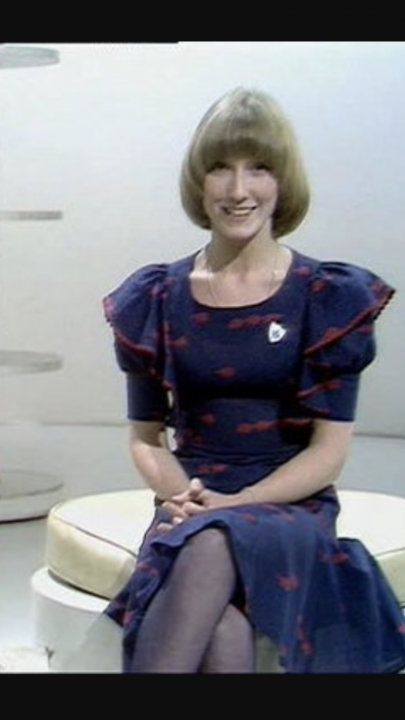 Weird Secret crush's from TV  - Page 14 - TV, Film & Radio - PistonHeads - The image is a low-quality photograph of a woman sitting on a beige chair against a white backdrop. The woman has short, straight blonde hair and is wearing a dark blue dress with a round neckline. She is smiling directly at the camera, and on her left cheek, there's a small, round emblem. The woman appears to be in a professional or formal setting, as indicated by the attire and background.