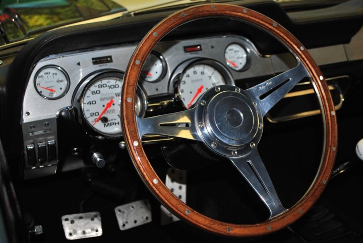 Pistonheads - The image captures a heartfelt scene from a classic car interior. The focus is on the steering wheel, which is set central to the vehicle's vintage aesthetic. Around the wheel are various gauges and dials, typical of older cars, all painted in a silver or chrome finish. The backdrop is mostly dark, highlighting the warm tones of the wood on the steering wheel and the synthetic leather of the vehicle's interior. The image seems to be a snapshot of a moment before hitting the road, the anticipation possibly felt by the car's owner.