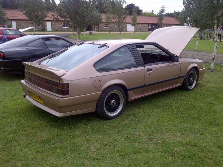 Pistonheads - The image features a vintage car exhibiting a convertible top that is partially open, revealing the trunk area. The car is parked on a grassy surface, suggesting an outdoor setting, possibly during an auto show or a personal collection event. In the background, there are buildings that appear to be part of a residential or commercial complex, indicating that the car is parked on or near a paved area. The car is painted in a light beige or light brown color, and the vintage nature of its design can be seen throughout its body and wheel rims.