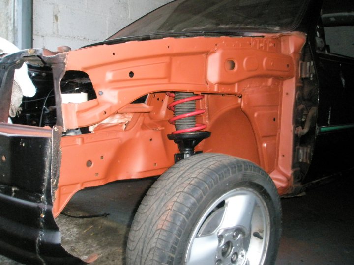 Fiesta Turbo Restoration Pistonheads - The image shows an orange body trabando on a vehicle chassis, with the focus on the suspension area. The red springs under the front end are exceptionally prominent, indicating a point that requires attention or is a problem area. The setting is indoors, possibly a workshop, given the presence of the car's uncorrected body amidst its skeletal structure. The front wheel of the vehicle is visible, engulfed in the orange material of the car's body. The car's interior is not visible, emphasizing the mechanical components of the vehicle.