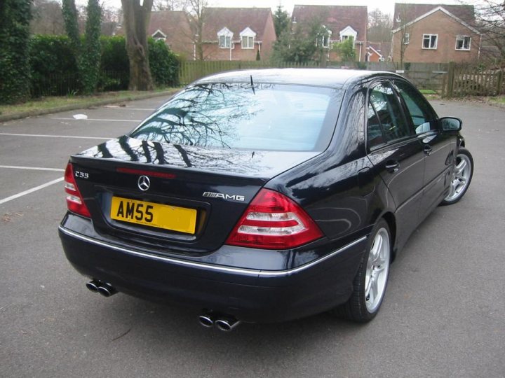 Pistonheads Amg Owner - The image shows a black Mercedes-Benz sedan parked in a parking lot. The car is gleaming and features a yellow license plate with the letters "AMS" indicating it likely belongs to a driver named Alex M. The car is equipped with a sunroof and the trunk lid is open, suggesting that it is a hatchback. The setting is peaceful with other cars parked in the background and greenery visible at the top of the frame.