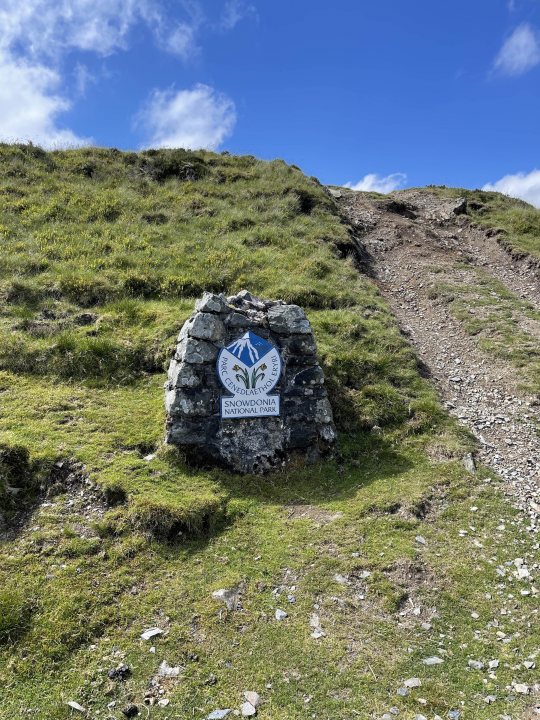 The "Photos From Today's Ride" thread. (Vol. 2) - Page 26 - Pedal Powered - PistonHeads UK - This image depicts a serene outdoor scene. At the center, there is a sign on a small structure or marker, possibly made of stone, with a logo above and text below it. The structure is situated on a grassy hillside, surrounded by wild vegetation and a few scattered rocks. The surrounding area appears to be quite rugged, with rocky terrain extending into the distance where more hills can be seen under a clear sky. There's no visible text that provides information about the location or signage in this image.