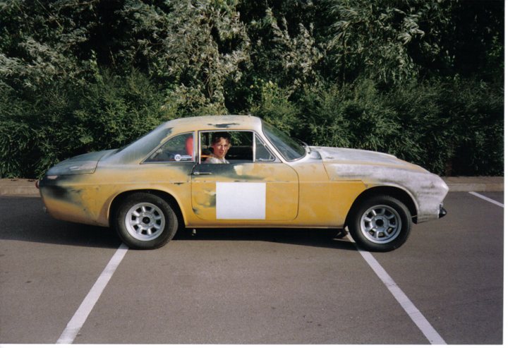 Scimitar Pistonheads Reliant - In the image, there's a scene set in a parking lot. A man is seen driving a yellow sports car. The car has a distinct white stripe running down its side and a glossy car paint that catches the eye. The man appears to be turning his head to the left, possibly eyeing the curb he's passing by. The setting is outdoors, under a clear sky. Near the left side of the image, there's a line of trees, providing a natural backdrop to the scene.