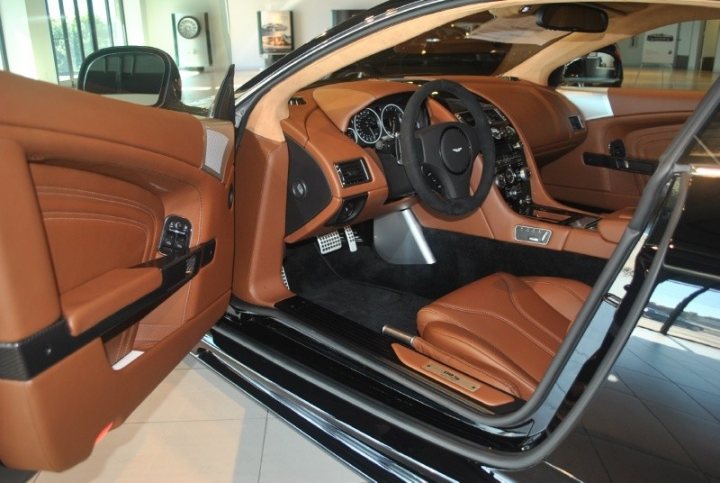 Sahara Tan Interior on an Aston - Page 1 - Aston Martin - PistonHeads - The image shows the impressive interior of a luxury car, featuring brown leather upholstery on both the driver's and passenger's side. In the foreground, the driver's side door is partially open, allowing a view along the length of the car. The car is equipped with state-of-the-art technology, evident in the high-quality infotainment system in the center console. The dashboard houses a variety of gauges and controls, indicative of a premium vehicle. The car is set against a backdrop that includes windows, suggesting the setting might be a well-lit showroom or a dealership. The attention to detail in the interior and the overall aesthetic exude a sense of refinement and elegance.