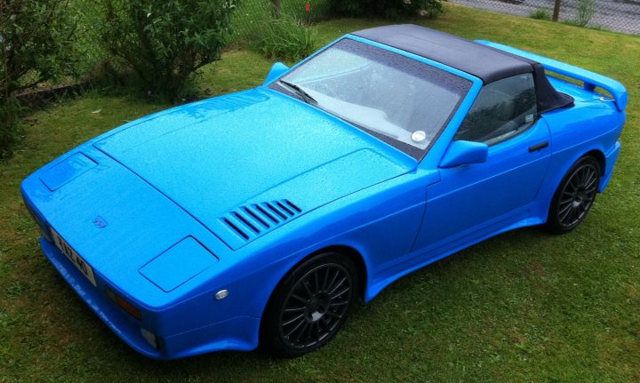 A blue car parked in a parking lot - Pistonheads - The image shows a vibrant blue, two-seater sports car parked on a grassy area. The car's sleek design and black soft top with two windows lend it a sporty and elegant appearance. It's equipped with black rims and a prominent rear spoiler. The background includes bushes, indicating a possible garden or park setting. The car's position, with its hood facing upward, suggests it's parked at an angle.