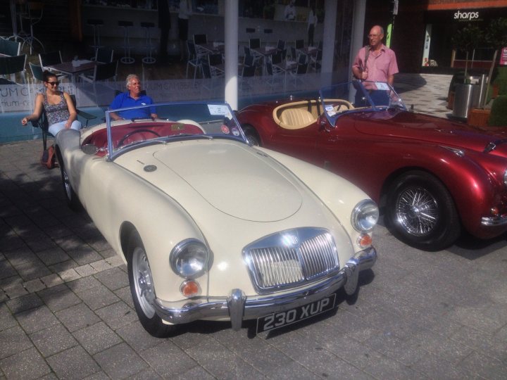 Camberley Town Centre car show - Page 1 - Thames Valley & Surrey - PistonHeads - This image depicts a vintage city street scene. Two sports cars are parked side by side: a classic red convertible and a vintage white roadster. They are sleek and shiny, reflecting the sunlight and giving a sense of their prestige and elegance. There are two individuals in the image; on the left, a woman in sunglasses is seated in the roadster, while on the right, a man is standing next to the convertible, appearing to admire the cars. The cars are parked in front of a shop, adding to the urban ambiance of the picture.