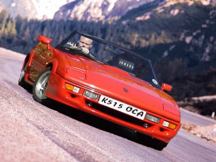 Sotw Pistonheads Reliant Scimitar - The image depicts a man driving an old, dark red sports car. He appears to be on a mountainous road, with a scenic backdrop featuring mountains. The car is speeding along the road, with the driver leaning forward to navigate the terrain. The license plate of the car is clearly visible.