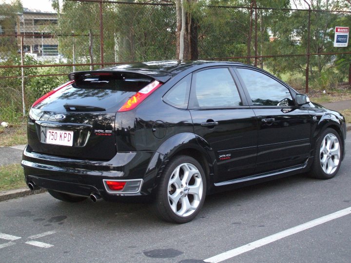 Spec Pistonheads Newbie Mazdaspeed Current - The image shows a parked black Ford Focus car. The vehicle is positioned on the side of a road or street, with the rear end facing the camera. The Ford logo is visible on the back of the car, as well as a red license plate. There's a design element near the taillights. The car is stationed behind what appears to be a wire fence, with greenery on the other side, indicating a residential or semi-urban area. A parking notice or sign is partially visible in the background, adding context to the setting.