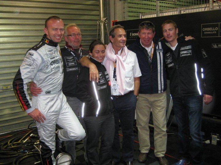 Merci les Belges/KRONOS - Page 1 - Aston Martin - PistonHeads - The image showcases a group of people in a garage setting, posing together for a photograph. In the foreground, there is a man in a white racing outfit seated and leaning towards two individuals in the back, who appear to be standing or slightly bending. The group includes a mix of men and women, with some individuals wearing lanyards around their necks that suggest they may be racing team personnel or event staff. The setting appears to be a workshop or garage area typically associated with motorsports, given the presence of sports cars in the background.