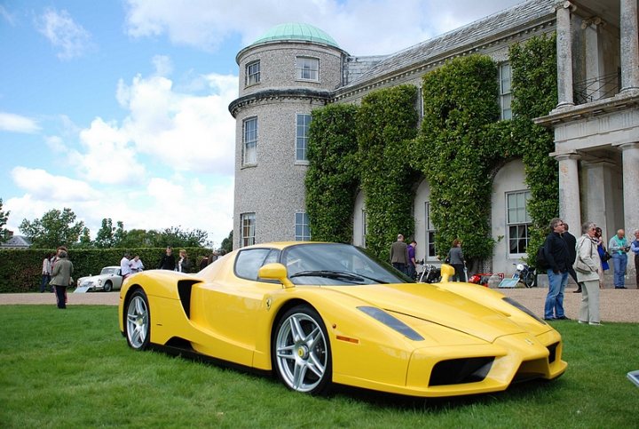 GRRC Lunch Club - Page 1 - Goodwood Events - PistonHeads - The image showcases a vibrant, long yellow sports car parked prominently on a lush green lawn. The car's sleek lines and shiny exterior indicate it is a Ferrari, a type of car renowned for its speed and luxury. The car is a convertible, with a visible top that is folded down. It occupies a large portion of the frame, indicating it is in the foreground. The setting suggests a special event or exhibition, with several people scattered around the perimeter of the car, possibly observing or admiring the vehicle. The car is in front of an old building with a green roof and greenery, contributing a contrast in architecture and style.