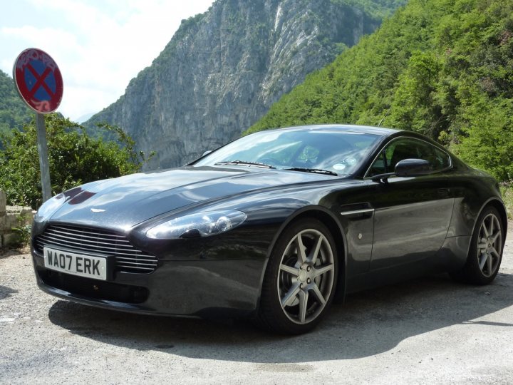 Trip of a Lifetime (picture heavy) - Page 1 - Aston Martin - PistonHeads - The image features a silver Aston Martin sports car parked on a road by the side of a green hillside. The car is oriented towards the camera and is set against a backdrop of a beautiful mountainous landscape. The surroundings include lush green trees and the hint of another road or highway. The day appears to be clear and bright, with the sky visible in the background.