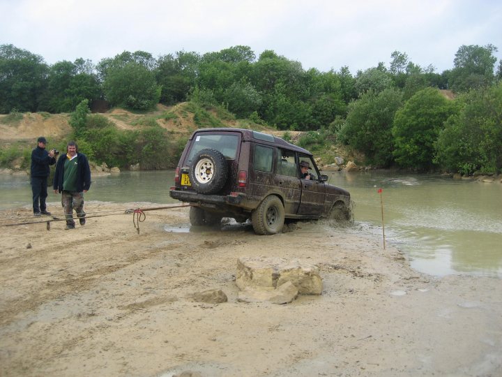 Champion V's Winchmax - Page 1 - Off Road - PistonHeads - In the image, an off-road vehicle, which appears to be a Land Rover, is partially immersed in muddy water, surrounded by a couple of individuals. The vehicle has large black tires and tinted windows. The two people are standing on the bank of the water, seemingly observing or possibly recovering from an incident involving the vehicle. For additional context, the people are wearing casual clothes appropriate for an outdoor activity, and to their left, the terrain is dry and sandy, indicating that the area around the water is quite different from where the vehicle is currently located.