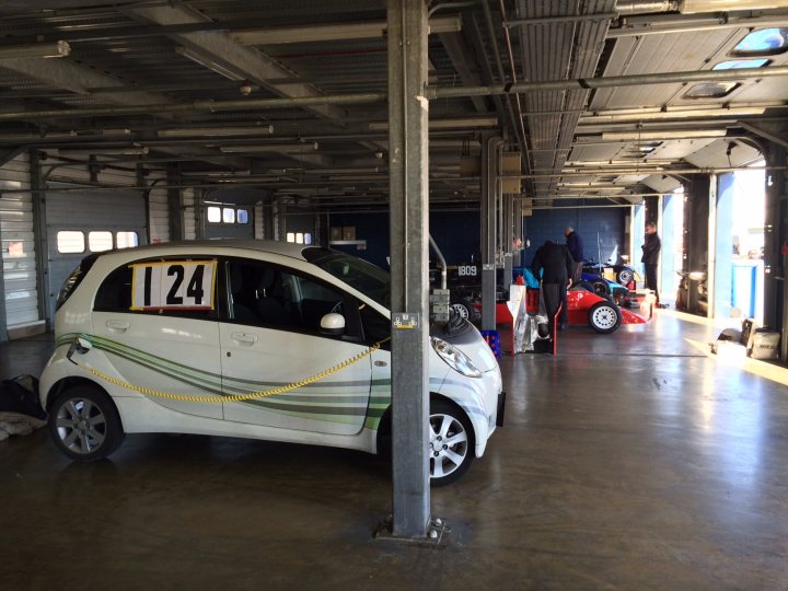 IMiev, Ion and C Zero - the forgotten electric cars - Page 1 - EV and Alternative Fuels - PistonHeads - The image shows a scene inside a large warehouse or garage, which is predominantly metal and well-lit. The primary focus is a small car, with its registration number prominently displayed on the back: "124." Next to the car, a motorcyclist is standing, accompanied by some people who appear to be inspecting or admiring the room and the machinery. In the background, there's a glimpse of additional mechanized items, and the floor is seen reflecting the high ceiling and the various industrial elements within the space.