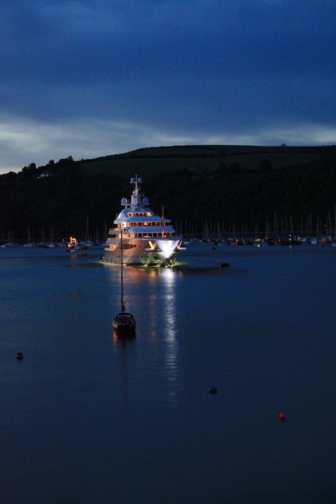 A small boat in a body of water - The image features a large luxury yacht situated at anchor in a body of water. The yacht's bright lights are on at night, providing a distinct contrast to its surroundings. The sky overhead is a deep navy blue, indicating that it is evening. The calm water reflects the yacht's lights based on its close proximity to the yacht. The water's surface is mostly still, adding to the serene night-time ambiance.