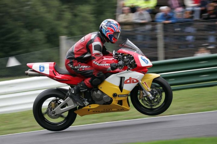 Pistonheads Notice Cbrrr Swapping - The image shows a professional motorcycle racer in the midst of a high-speed run on a track. The rider is dressed in full racing gear, indicating the intensity of the race. The motorcycle is highly aerodynamic, designed for speed, and is in a dynamic pose, leaning to the right as if in the midst of a turn. The track appears to be an outdoor venue, and in the background, spectators can be seen watching the event from what looks like an infield viewing area. The overall feeling of the image is one of motion and high performance, capturing the essence of motorcycle racing.