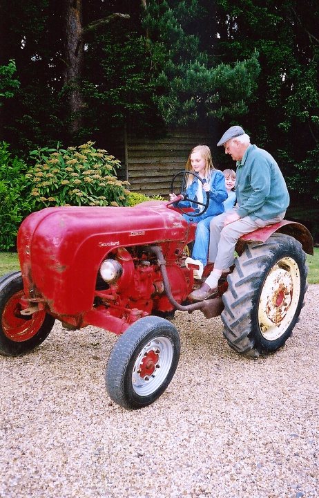 Tribute To A Racer - Page 1 - Classic Cars and Yesterday's Heroes - PistonHeads - The image shows a man and a young girl riding on the back of a vintage red tractor. The tractor is an older model, possibly vintage, with a classic red body and black tires. It's parked on gravel, and the background reveals a lush green landscape with trees and shrubs. The man appears to be an older individual, dressed casually, and the girl is seated in front of him, looking forward with a slight smile. The scene seems to suggest a day out in the countryside or on a farm, where the ride on the tractor is being enjoyed.