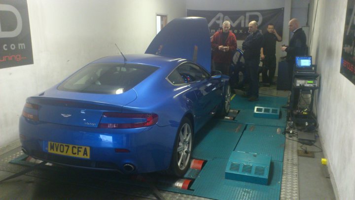 Arggh Clutch - Let the mods begin! - Page 1 - Aston Martin - PistonHeads - The image shows a blue sports car inside a garage area. A group of men, clad in jackets, are inspecting the car. The car's hood is open, revealing the engine for examination. The garage floor is made of white tiles, and safety precautions appear to be in place, such as a pallet under the back wheels of the vehicle. Various diagrams or images seem to be in the background, possibly related to automotive services or repair.