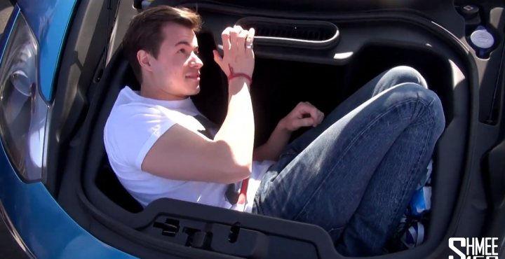 A woman sitting in a car holding a cell phone - Pistonheads - The image depicts a young man sitting in the trunk of a sports car. He is casually dressed in a t-shirt and jeans, with brunette hair and a beard. His hands are holding something pen-shaped, possibly taking a selfie or looking at his phone. The interior of the car is dark, contrasting with his lighter attire. The scene suggests a candid, relaxed moment captured within the confines of a convertible vehicle.