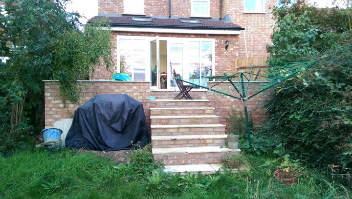 Planning permission for a patio - Page 1 - Homes, Gardens and DIY - PistonHeads - The image shows an outdoor setting consisting of a residential brick building with a ground-level window and a garage door. Tall, overgrown plants and bushes are obscuring both the bottom of the building and some of the garden area in front of it. On the left side of the house, a covered patio area can be seen, featuring a green vegetable trellis and an outdoor dining table with lawn chairs. There is a covered stairway leading up to the patio area. To the left and right of the flight of stairs, there are potted plants. A flip-flop is visible on the left stairs. The sky is visible in the background, suggesting it could be early morning or late afternoon given the soft, even lighting.