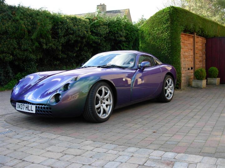 Pistonheads Tuscans Reflexflip Paint - The image shows a unique purple sports car parked on a driveway. It's positioned in front of a cohesion-style hedge that forms the backdrop. The car is equipped with a large air intake near the front and has a low-slung profile. Its design and paint suggest it could be a handcrafted or custom-built vehicle. The setting indicates a residential area with paved driveways and landscaped hedges.