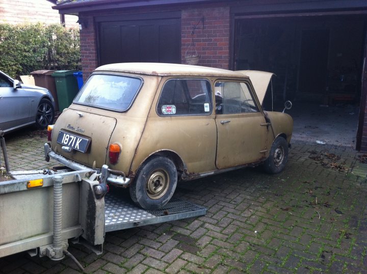 My 1960 Austin seven - Page 2 - Classic Minis - PistonHeads - This image depicts an old, brown Mini Cooper car that is being pulled by a tow car. The tow car is positioned in a driveway with a brick surface and appears to be a professional towing service. The Mini Cooper is positioned at an angle, with its front lifted off the ground. The car has a European license plate and is situated in front of a garage or shed.