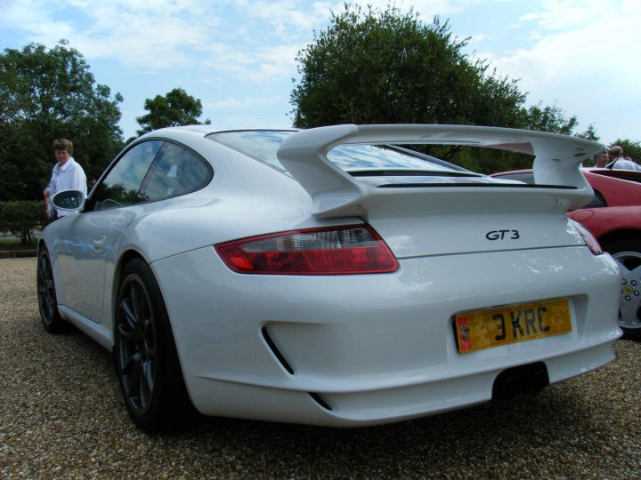 Breakfast Meeting, The Hare, Roxwell 4th June - Page 1 - Kent & Essex - PistonHeads - The image features a new white Porsche GT3 with the license plate reading "3KRC" on a clear day. The car is parked on a gravel area with grass and trees in the background. The tire pattern appears to be a testament to the car's sporty nature. In the background, there are trees and a person walking away from the camera. There is a second car, a red one, slightly out of frame to the left. The car's sleek design and imposing presence are the focal points of the image. The overall scene suggests a quiet and sunny day in a park or a similar open space.